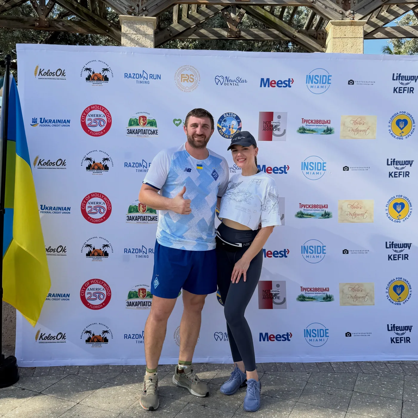 Two participants in front of the sponsor wall at the Razom Freedom Run, next to a Ukrainian flag