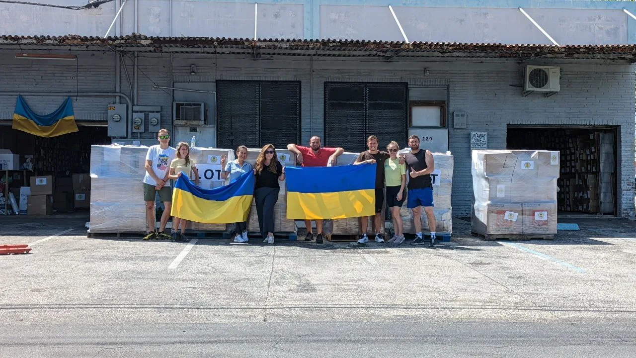 FL4UA volunteers holding Ukrainian flags in front of pallets of aid at the Hallandale warehouse