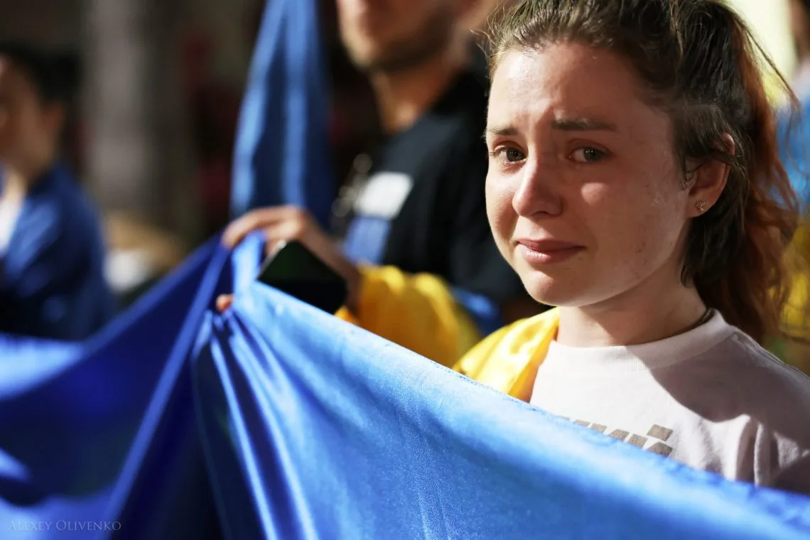 A woman in tears holding a large blue and yellow flag at a community gathering