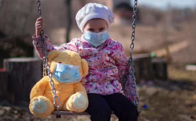 A child in a face mask sitting on a swing with a teddy bear